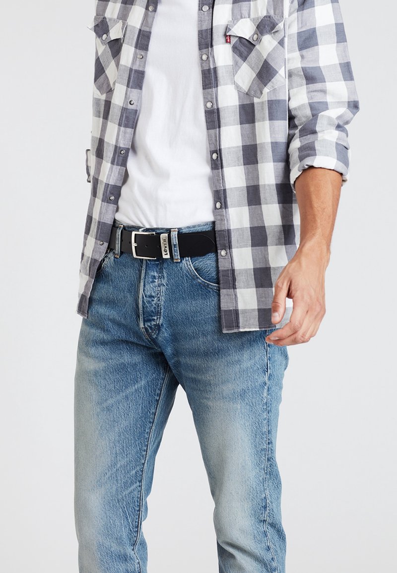 Homme portant un jean bleu avec une ceinture noire Levi's, un t-shirt blanc et une chemise ouverte à carreaux gris et blanc aux manches retroussées.