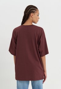 Bordeaux oversized t-skjorte med korte ermer, rund hals og glatt bomullsstoff. Modellen har på seg lyseblå denimjeans.