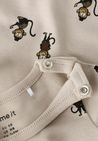 Plongée en coton beige avec des singes en dessin animé imprimés en marron et noir. Présente un col rond et des fermetures à pression à l'épaule.