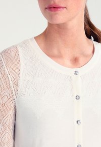 Vit kofta med texturerade spetsärmar, rund halsringning och fem dekorativa knappar. Slät tyg med ett delikat mönster.