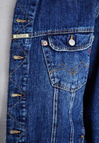 Denim jacket in blue with yellow stitching, featuring a chest pocket, button closure, and a "REPLAY" label. Notable zigzag stitching detail.