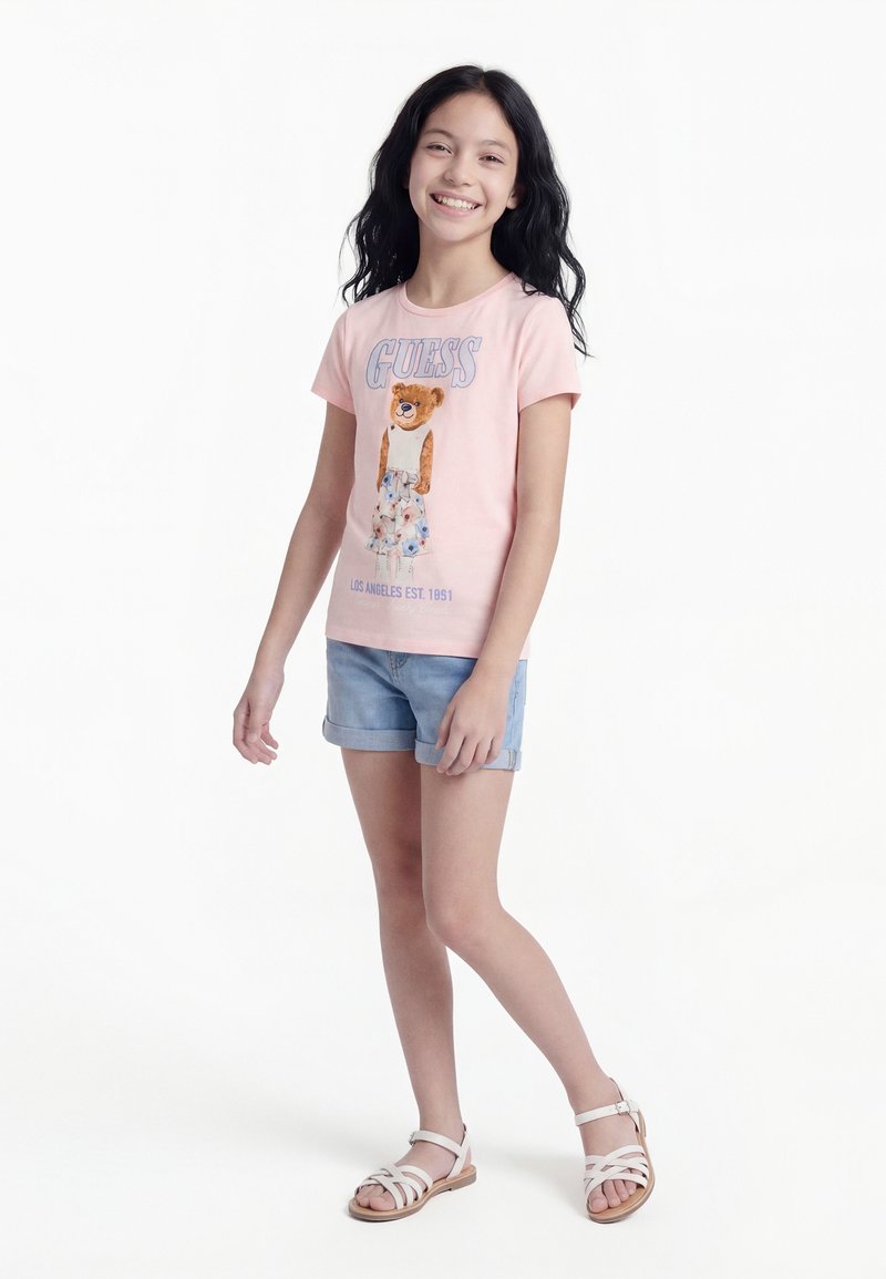 Κορίτσι με μακριά σκούρα μαλλιά που χαμογελά, φορώντας ροζ μπλούζα με στάμπα αρκουδάκι, τζιν σορτς και λευκά σανδάλια, στέκεται πάνω σε λευκό φόντο.
