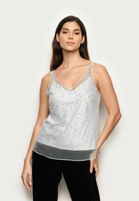 Przezroczysta biała koszulka na ramiączkach z dekoltem w serek, ozdobiona metalowymi ćwiekami, z regulowanymi ramiączkami i swobodnym krojem. Pięciowarstwowy design u dołu.