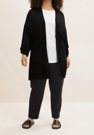 Cardigan kimono léger noir avec des manches trois-quarts, devant ouvert, associé à un haut blanc, un pantalon noir et des sandales tissées noires.