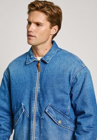 Chaqueta de mezclilla en azul claro con cierre de cremallera en la parte delantera, cuello puntiagudo y dos bolsillos de solapa con botón. Presenta detalles de costura y un corte relajado.