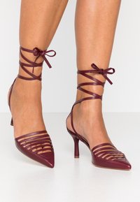 Sandali con tacco alto in pelle bordeaux, con punta affusolata, design con cinturini e lacci alla caviglia. Texture liscia con dettagli a fessura sulla parte superiore.