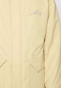 Veste beige clair avec poches à rabat et petit motif de flamme brodé sur la partie supérieure gauche de la poitrine.