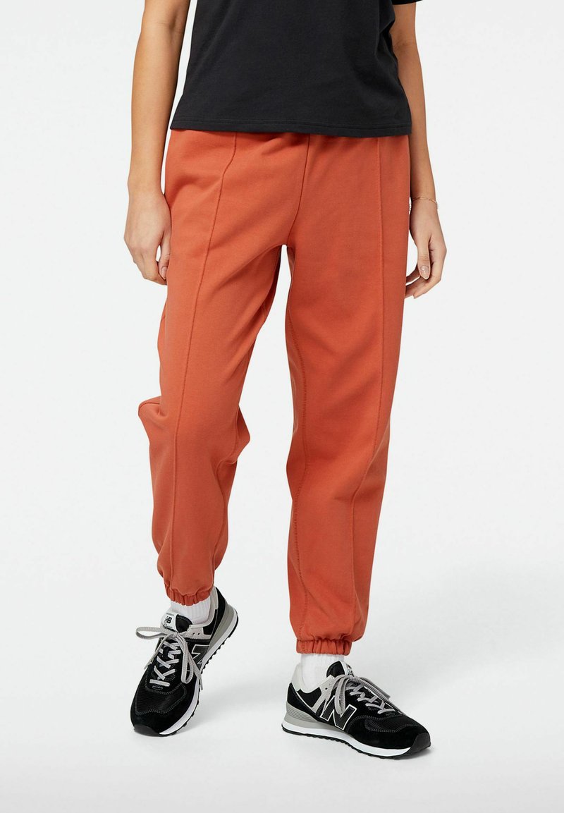 Orange Sweatpants mit elastischen Bündchen und Seitennähten, kombiniert mit schwarzen Sneakers, die weiße und graue Akzente auf einem schlichten Hintergrund aufweisen.