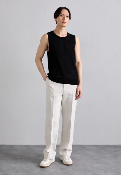 Black sleeveless ribbed top, white tailored trousers, and light athletic shoes. Model stands with one hand in pocket against a neutral background.