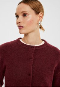 Mörkröd cardigan med rund hals, med tre knappar fram, tillverkad av mjukt, texturerat tyg. Vit skjorta under.