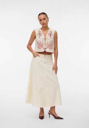 Femme portant un gilet sans manches crème avec une broderie de feuilles roses, associée à une jupe longue évasée crème et des talons sombres à bout pointu.