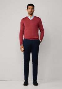 Mężczyzna stojący w czerwonym swetrze w serek na jasnoniebieskiej koszuli, granatowych spodniach oraz ciemnych butach na sznurówki, na jednolitym tle.