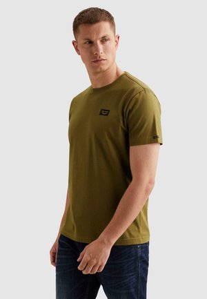 Homme portant un t-shirt à manches courtes vert olive avec un petit logo noir sur la poitrine et la manche, associé à un jean bleu foncé, regardant de côté.