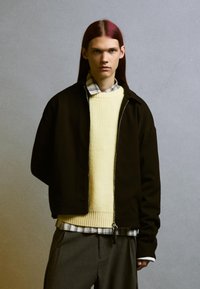 Fekete cipzáras dzseki egy sárga kötött pulóver felett, kockás inggel rétegezve. Szürke csíkos nadrágok teszik teljessé a megjelenést.