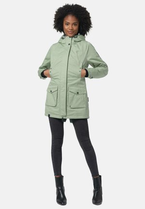 Parka légère verte avec une fermeture éclair à l'avant, deux grandes poches avant, une capuche ajustable et des accents noirs sur les manches. Associée à un jean foncé.