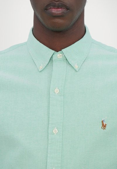Chemise à boutons vert clair en tissu texturé. Dotée de boutons blancs et d'un petit logo brodé représentant un joueur de polo à cheval.