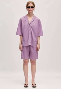 Lila oversized knoopoverhemd, bijpassende shorts en zwarte sandalen. Gladde stof, relaxed pasvorm, korte mouwen en minimalistisch ontwerp.