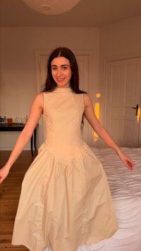 Νεαρή γυναίκα που χαμογελά, φοράει μπεζ αμάνικο φόρεμα, στέκεται σε ένα ζεστά φωτισμένο υπνοδωμάτιο με ξύλινο πάτωμα και λευκό κρεβάτι.