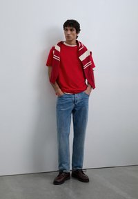 Jonge man staat tegen een witte muur, draagt een rood T-shirt, een gestreepte trui op de schouders, lichtblauwe spijkerbroek en donkerbruine schoenen.