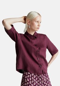 Donna bionda che indossa una camicia a bottoni viola scuro e ampia, con una gonna a fantasia, con una mano all'anca e l'altra dietro la testa.