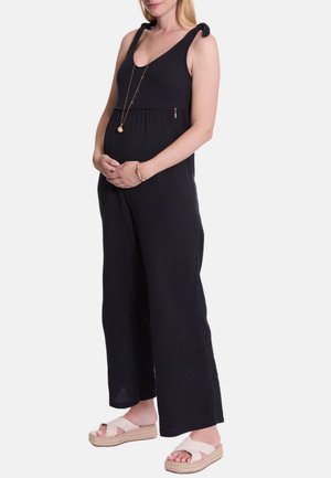 Zwarte mouwloze jumpsuit met strikbanden en een ontspannen pasvorm, met een geplooide naad boven de taille. Gedirigeerd met lichtgekleurde sandalen.
