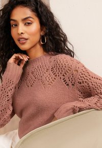 Strickpullover in zartem Rosé mit einem komplexen Spitzenmuster auf den Schultern. Mit langen Ärmeln und einem gerippten Kragen.