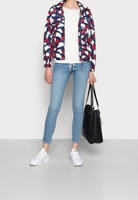 Kleurrijke windjack met een abstract patroon in rood, wit en blauw, gecombineerd met een wit t-shirt en lichte blauwe skinny jeans, en een zwarte handtas.
