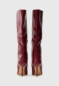 Un par de botas altas de cuero rojo oscuro con tacones altos y cremalleras laterales, vistas desde atrás sobre un fondo blanco liso.