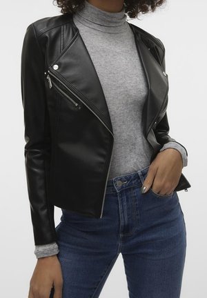Femme portant une veste en cuir noire par-dessus un col roulé gris et un jean bleu, main reposant dans la poche avant du jean.