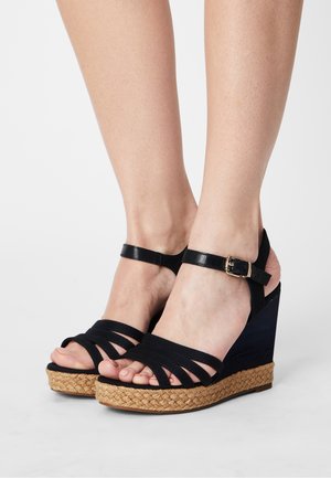 Sorte kilehæle med vævet jute hæl, stroppedesign og en ankelrem med spænde. Glat overmateriale og åben tå stil.
