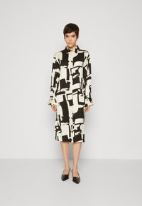Rochie cu mânecă lungă dintr-un material crem, cu un model abstract în alb și negru, având un guler și accente cu nasturi la manșete.