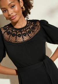 Robe noire avec un empiècement en dentelle florale, des manches bouffantes et une taille cintrée avec une ceinture assortie. Présente un tissu texturé et des détails complexes.