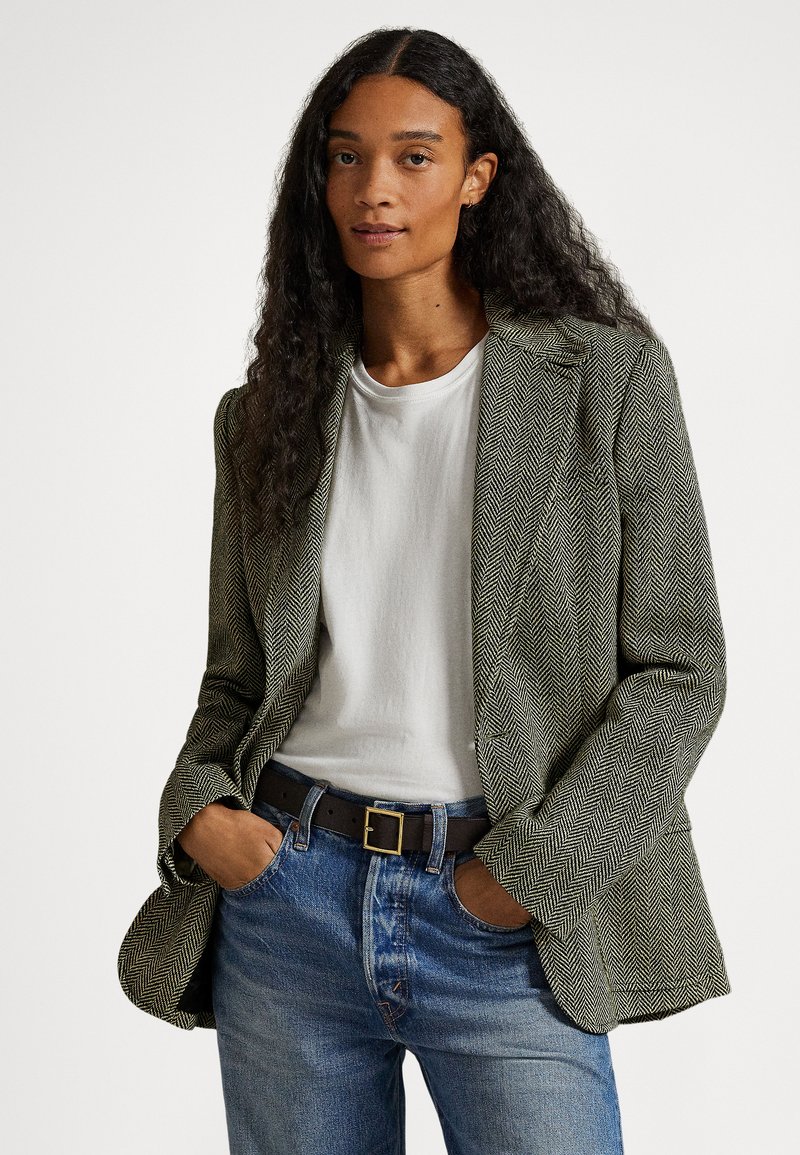 Kobieta z długimi kręconymi włosami, ubrana w białą koszulę, zieloną marynarkę w jodełkę, niebieskie dżinsy i czarny pasek, stoi z rękami w kieszeniach.