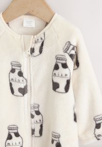 Grenouillère bébé en polaire couleur crème avec fermeture éclair, ornée d'un motif de biberons imprimé vache en gris et blanc. Texture douce.