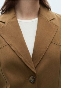 Femme portant un blazer beige à large revers par-dessus un haut blanc côtelé à col rond, mettant l'accent sur la poitrine et le cou.