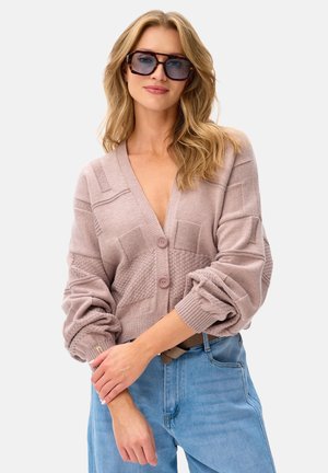 Kobieta o falowanych blond włosach, nosząca duże okulary przeciwsłoneczne w oprawkach w kolorze żółwia, jasnoróżowy, teksturowany sweter rozpinany oraz jasnoniebieskie dżinsy z wysokim stanem.