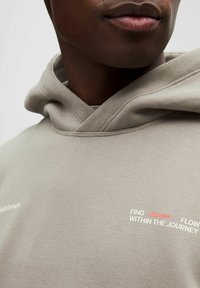 Haut à capuche beige avec une texture douce, présentant une capuche en croix et un texte graphique en blanc et rouge : "TROUVEZ LE COURANT À L'INTÉRIEUR DU VOYAGE."