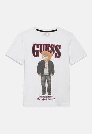 Valkoinen puuvillainen T-paita, jossa on kuvituskarhu mustassa takissa ja farkuissa, punainen "GUESS"-teksti yläpuolella ja logo alhaalla.