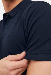 Marineblaues Polohemd aus strukturiertem Stoff, mit einer Knopfleiste mit zwei Knöpfen und kurzen Ärmeln, sowie einem kleinen Logo auf der Brust.