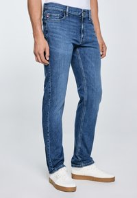 Férfi kék slim-fit farmerben és fehér tornacipőben, egyszínű világos háttér előtt állva.