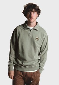 Sudadera verde oliva con cremallera de cuarto, con una tela texturizada y un pequeño parche de logo beige en el lado izquierdo del pecho. Pantalones marrones.