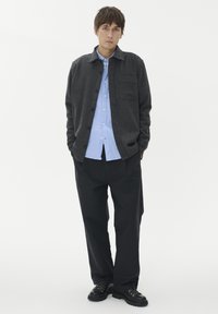 Oglasta srajčna jakna v ogljeni barvi preko svetlo modre srajce, kombinirana z ohlapnimi črnimi hlačami in črnimi čevlji. Jakna ima žep na prsih.