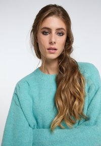 Un maglione lavorato a maglia di un azzurro chiaro con un collo rotondo e maniche leggermente a sbuffo, indossato da una persona con capelli lunghi e ondulati.