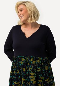 Robe à manches longues avec un haut marine et une jupe à motifs en vert, jaune et bleu, présentant un décolleté arrondi et une taille froncée.