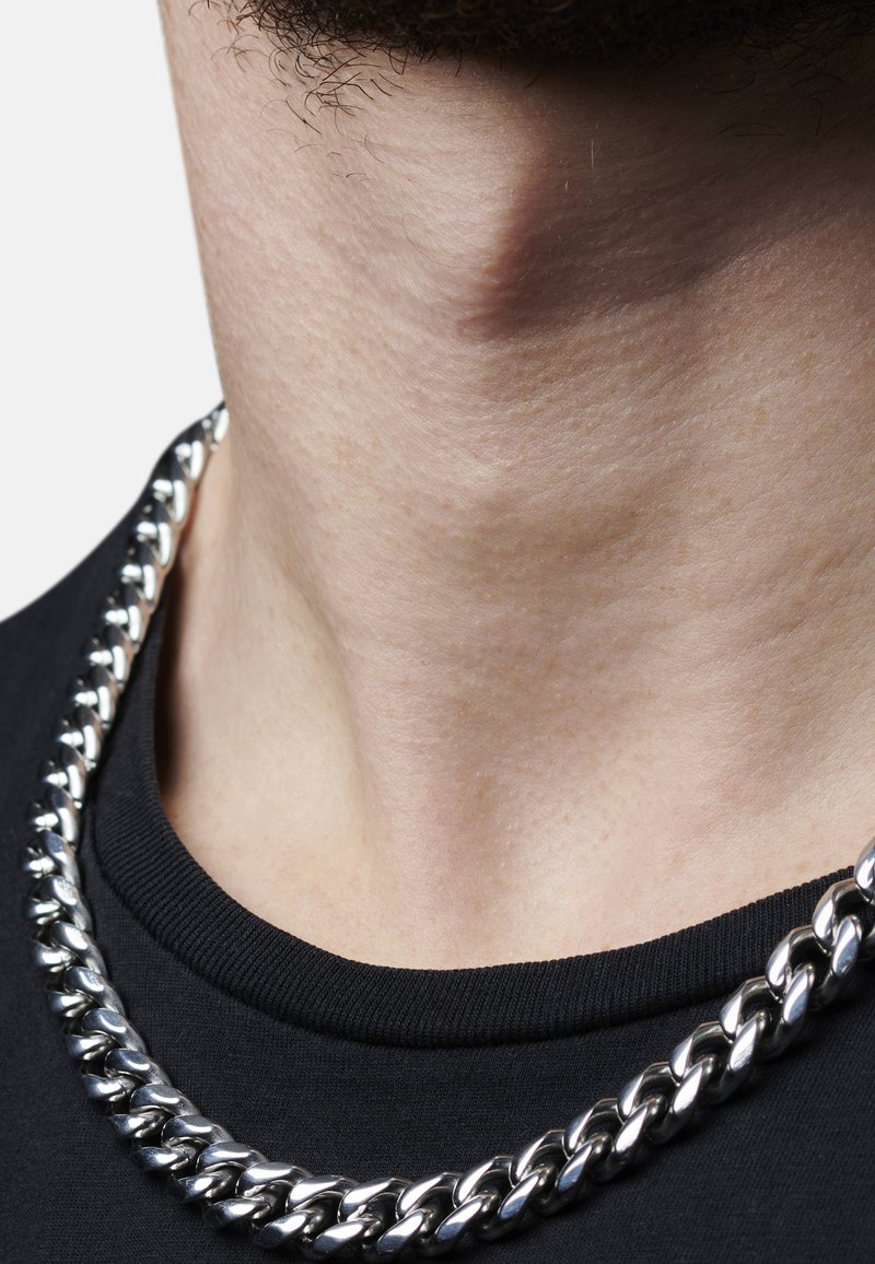 Sølv kædedesign med tykke led, båret over en sort t-shirt med rund hals. Glat tekstur med en skinnende metallisk finish.