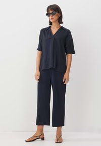 Marinefarbenes, kurzärmliges, kragenloses Oberteil kombiniert mit locker sitzenden, marinefarbenen Hosen. Das Model trägt schwarze Sonnenbrillen und dünne schwarze Sandalen. Minimalistisches Design.