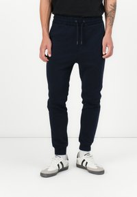 Marineblaue Jogginghosen mit elastischem Bund, gerippten Bündchen und glatter Textur, kombiniert mit weißen Sneakers mit schwarzen Streifen.