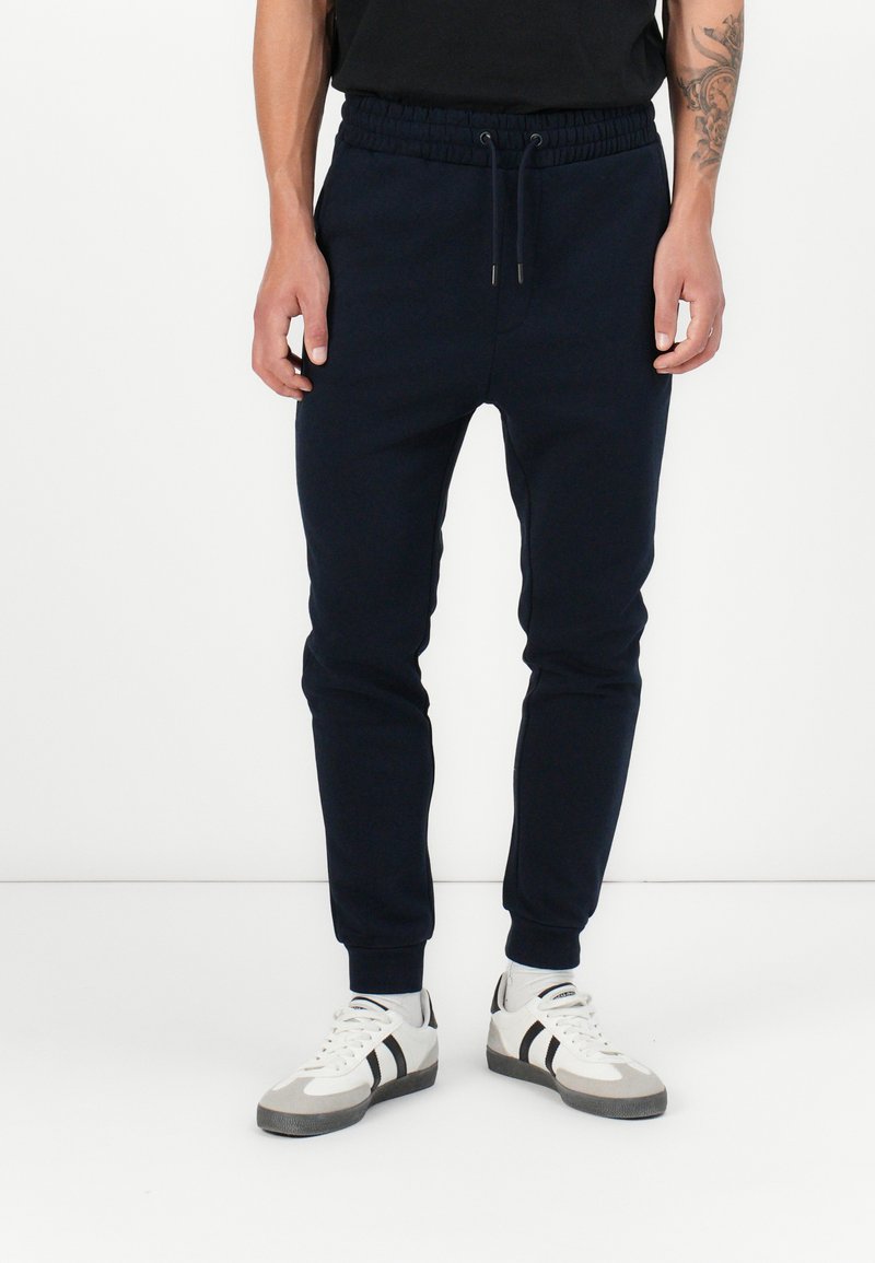 Marineblaue Jogginghosen mit elastischem Bund, gerippten Bündchen und glatter Textur, kombiniert mit weißen Sneakers mit schwarzen Streifen.