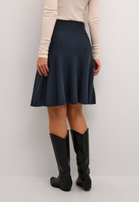 Ženska, oblečena v mornarsko modro razširjeno krilo, bež dolg rokav in črne škornje do kolen, stoji ob preprostem svetlem ozadju.