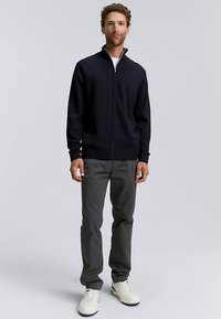 Mornarsko moder pulover z zadrgo in visokim ovratnikom, nošen čez belo srajco, kombiniran s sivimi hlačami in belimi supergami. Gladek pleteni vzorec.